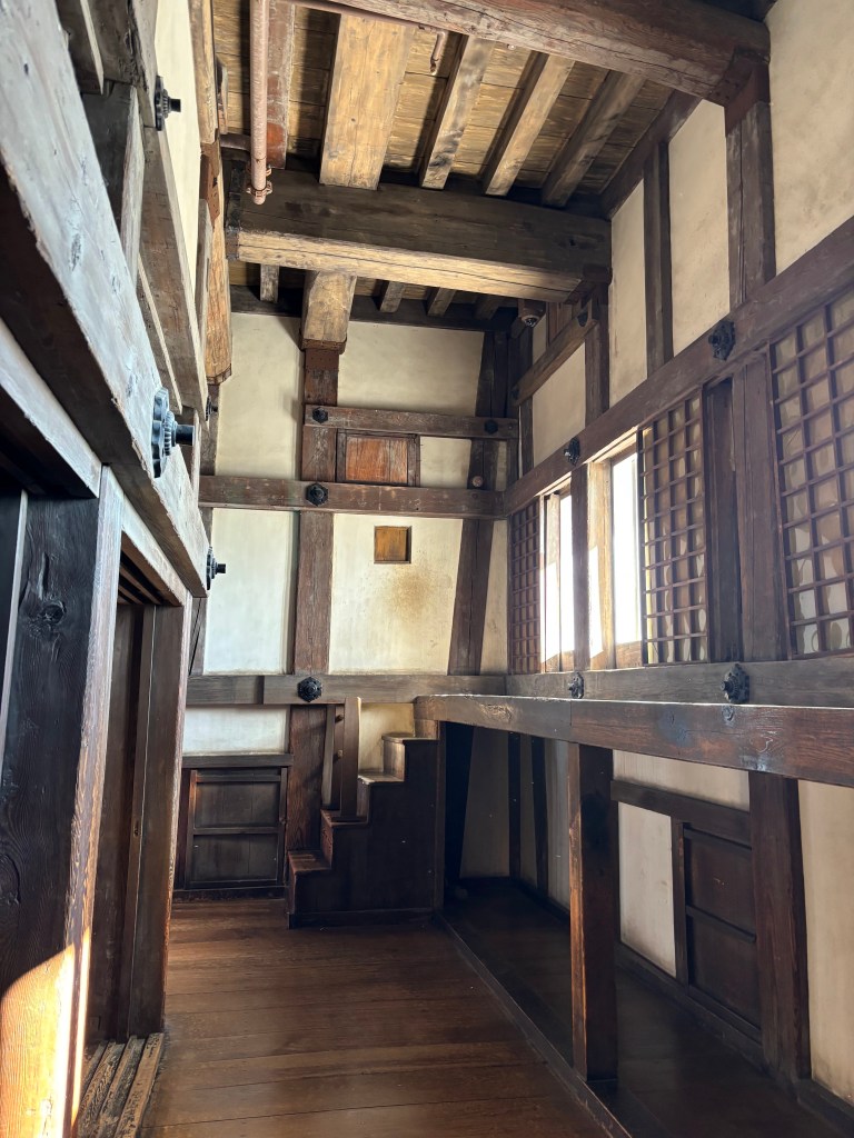 Interior corridor inside Himeji Castle with wooden beams, lattice windows, and steep wooden stairs.